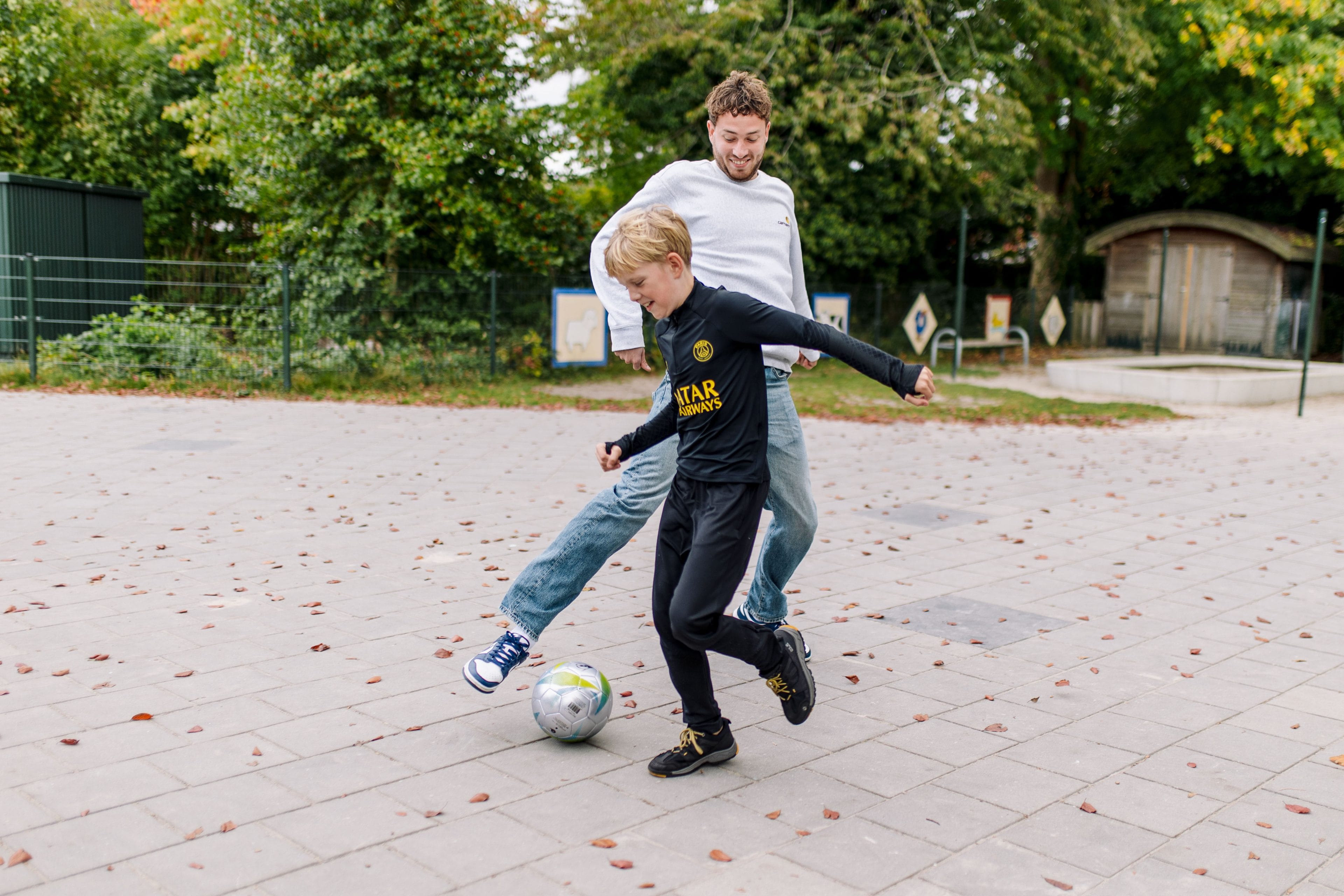 Begeleider en jongere samen aan het voetballen op een plein